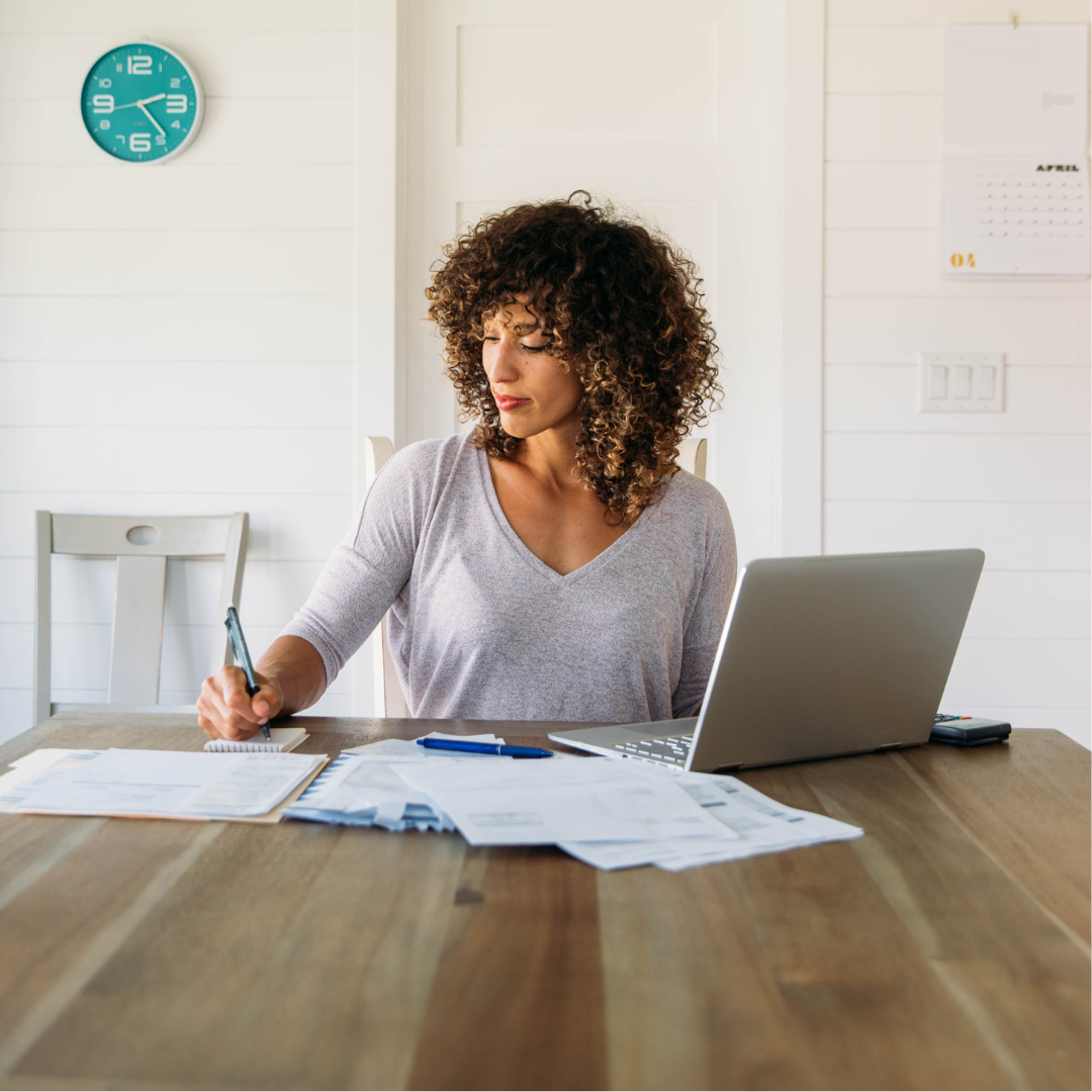 Woman writing on paperwork at a table with a laptop and financial documents