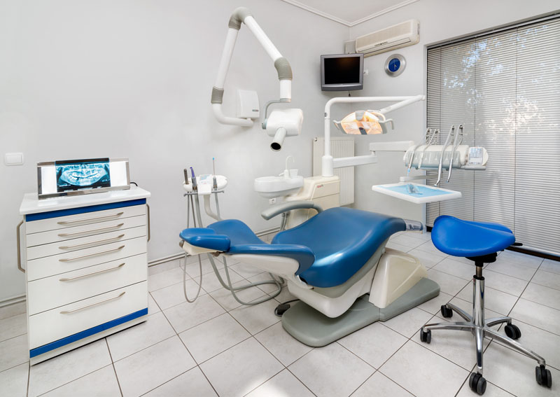 Modern dental treatment room with blue patient chair, exam light, X-ray monitor, and stool