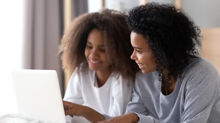 Two smiling young women with curly hair looking at a laptop screen together