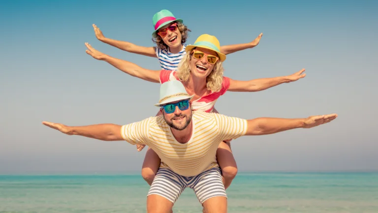 Smiling family stacked on dad's shoulders at the beach, arms outstretched, wearing sunglasses and hats