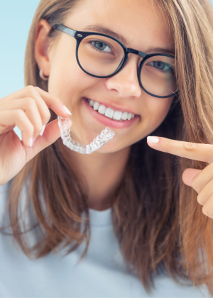 Smiling teen in glasses holding a clear aligner and pointing to her teeth